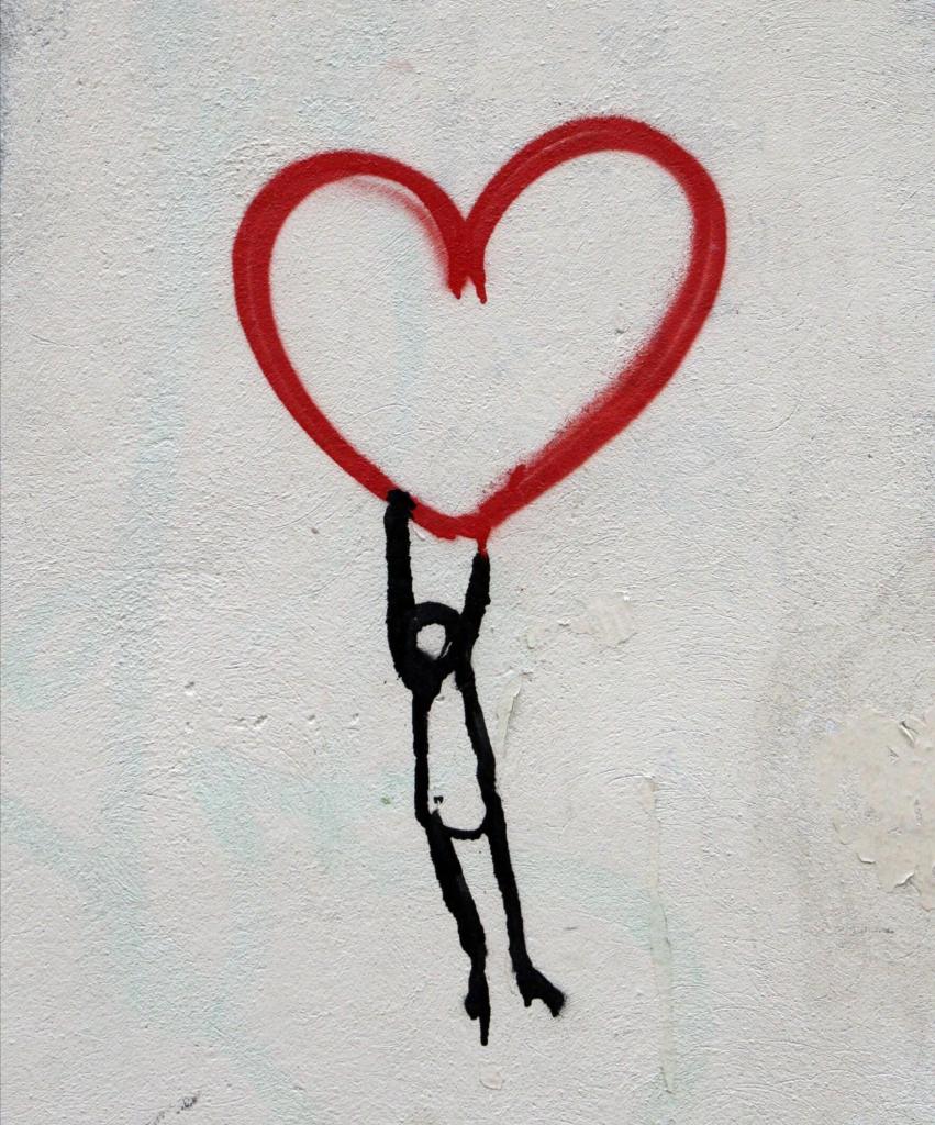 A white-washed external wall with a heart outline spray painted in red. A stick figure spray painted in black is shown hanging from the bottom of the heart.