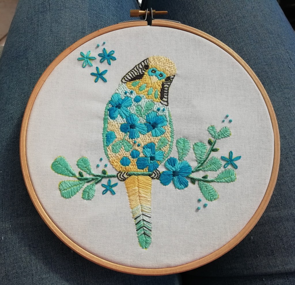A hand-stitched picture of a yellow budgie on a spring branch, displayed in an embroidery hoop.