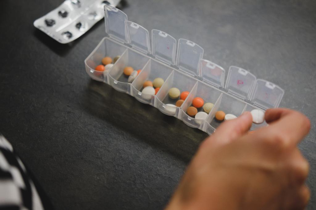 A hand in the process of putting tablets of different colours into a plastic dispenser/organiser with seven segments. An empty blister pack lies in the background.