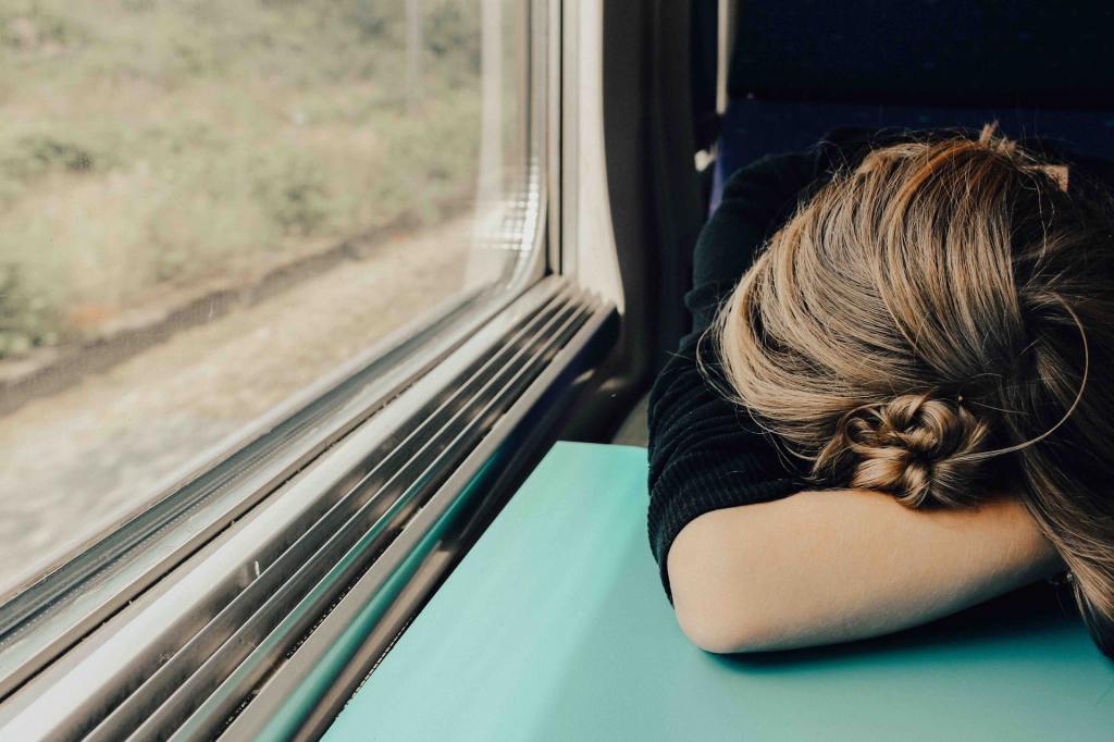 A young woman is hunched over a train table, resting her head on her arm, possibly asleep.