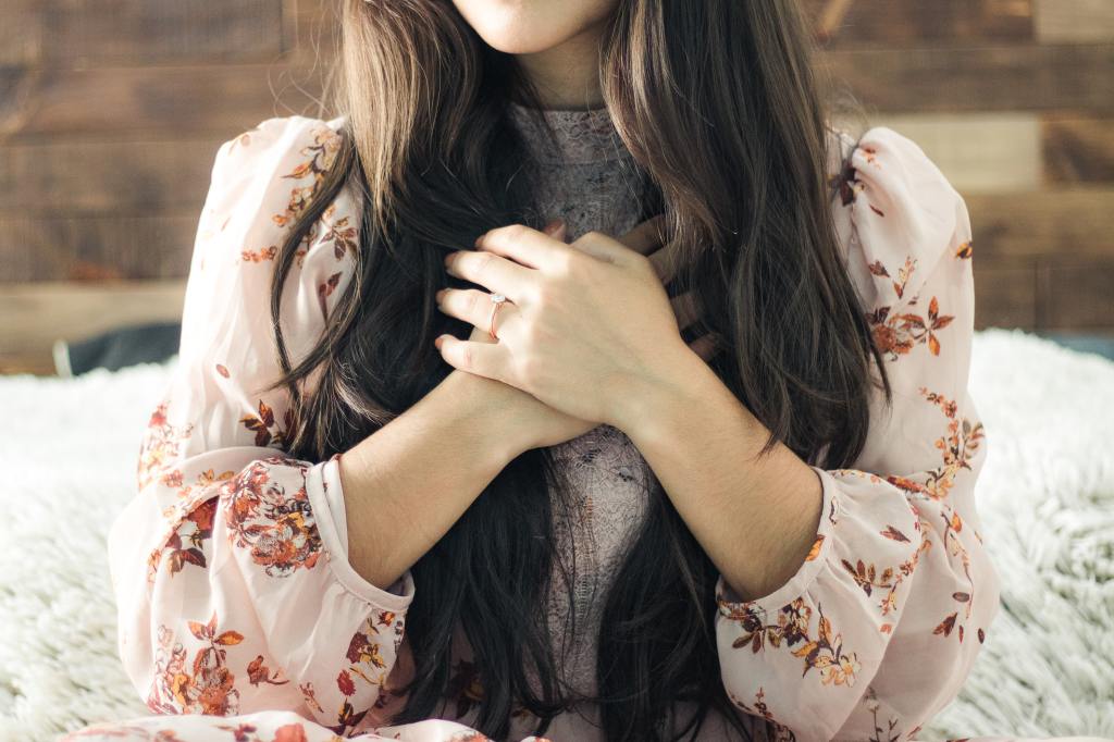 The torso and shoulders of a young woman with long brown hair wearing a floral shirt. She has her hands placed on her chest, one on top of the other.