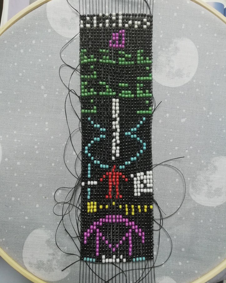 A representation of the Arecibo message, made from tiny beads woven together. Loose black threads protrude at various places and a grey fabric with stars and moons sits behind the piece.