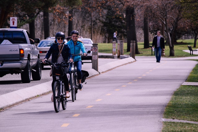 Two people cycling in a cycle lane. In the background is a park with people walking.