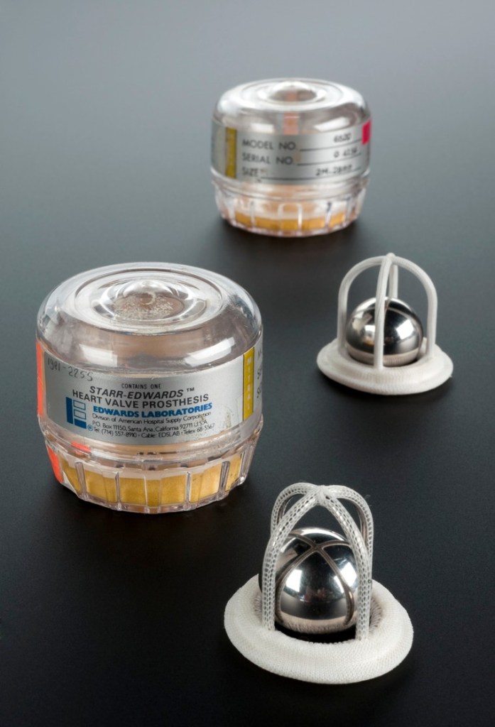 On a table sit two clear containers with labels reading, "Starr-Edwards heart valve prosthesis, Edwards Laboratories." The valves sit on the table - four-legged, open-sided domes of metal covered in mesh-like fabric. Inside each dome sits a close-fitting metal ball/ballbearing.