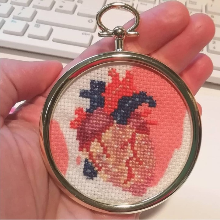 An anatomical image of a human heart, stitched onto aida fabric that is white with pale red blobs intended to look like blood stains. The cross-stitched heart is mounted in a small, round, gold coloured frame, which is held in the palm of a hand. A computer keyboard is visible in the background. 