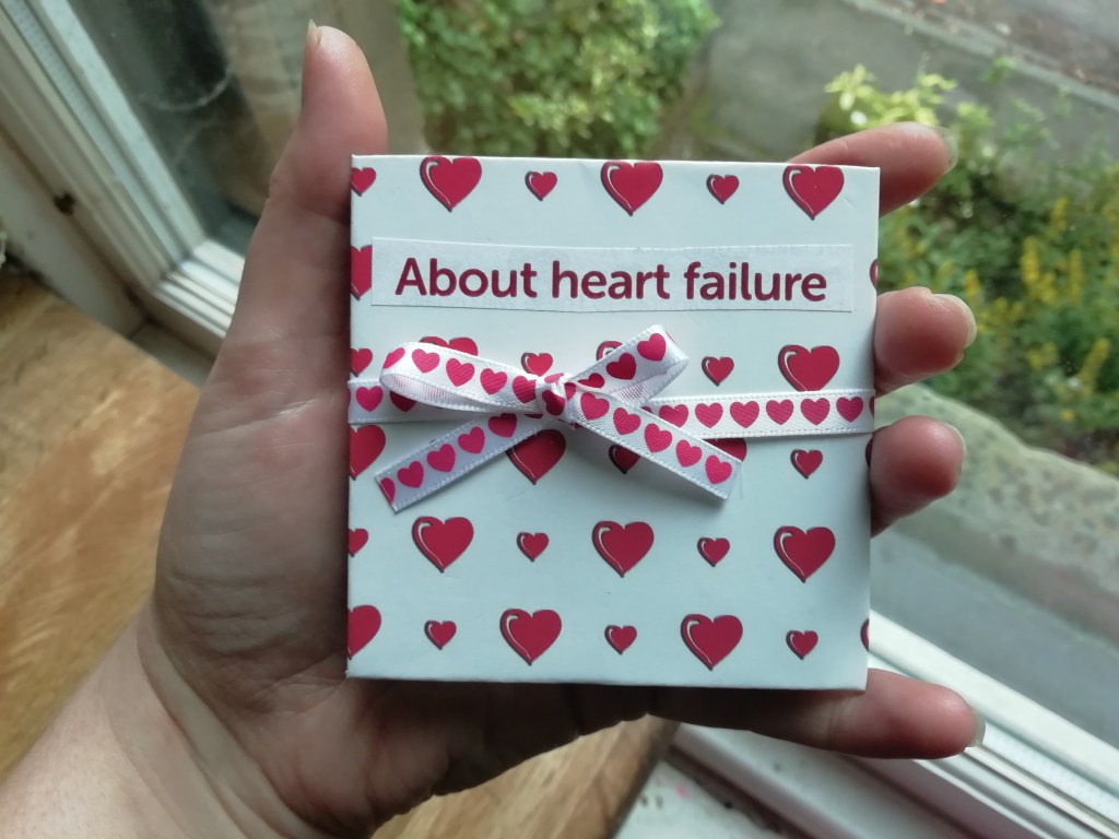 A hand holds a cardboard square approximately 8cm across. The square is covered in white paper with cartoon-style red love-hearts. Across the centre is a white ribbon with red hearts, tied in a bow. A title (cut from paper, with red text on a white background) reads "About heart failure."