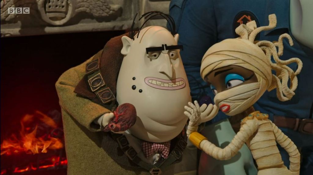 A still from stop-motion animated children's television programme Scream Street. Otto Sneer (a bald man with heavy dark eyebrows and multiple chins, wearing a mayoral chain) holds a realistic-looking heart in his hand, while Cleo (a sporty-looking Egyptian mummy, wrapped in bandages and wearing blue eye make-up) reaches out towards it. A fire blazes in a hearth in the background.