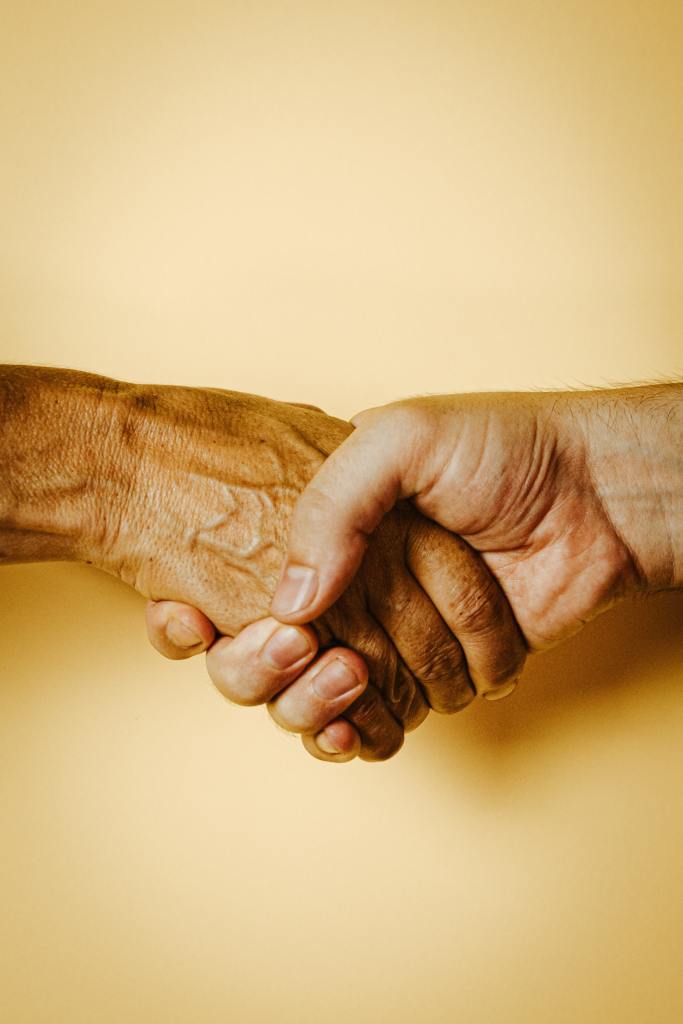 Two adult people's hands hold one another in a handshake-type grip. The photo is cropped at the wrists and has a plain pale yellow background.