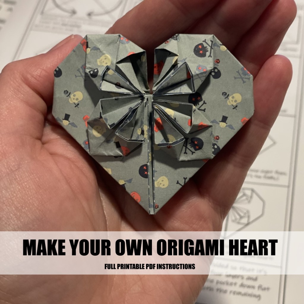 Photograph of a hand holding an origami heart. Text superimposed on image reads "Make your own origami heart, full printable PDF instructions."