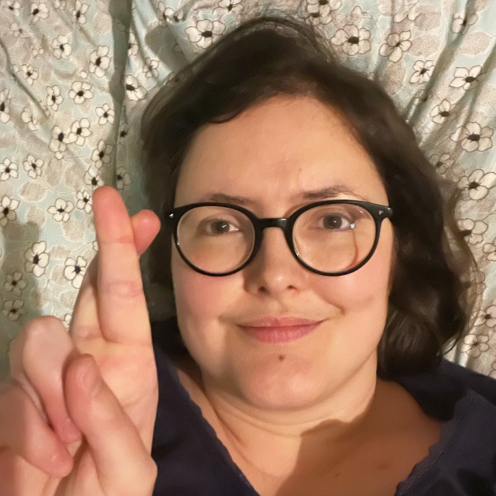 Selfie of Laura (white woman in her 40s with dark hair and dark rimmed glasses) lying in bed, slightly smiling. She holds her hand up in the foreground with her fingers crossed, forming an X shape.